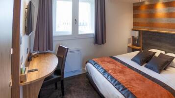 Ein Hotelzimmer mit Bett, Schreibtisch, Stuhl, einem Fenster mit Vorhängen und einem an der Wand befestigten Fernseher.
