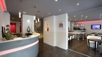 A modern hotel lobby with a reception desk, seating area, and a staircase leading to an upper level.
