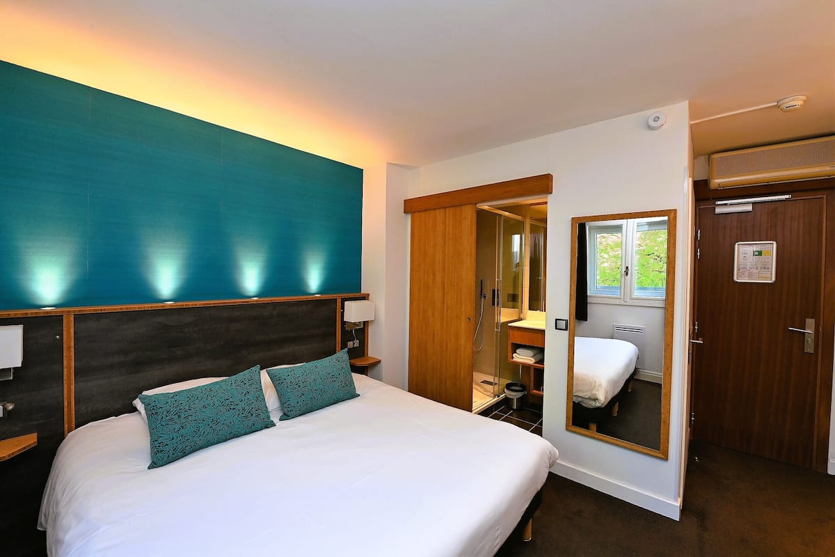 A hotel room with a large bed, a wooden headboard, a mirror, a wardrobe, and a small bathroom.