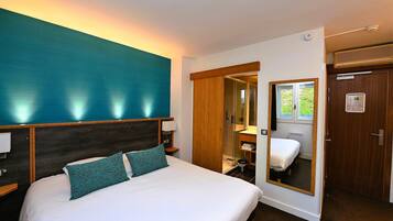 A hotel room with a large bed, a wooden headboard, a mirror, a wardrobe, and a small bathroom.