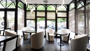 L’intérieur d’un café moderne, avec des tables rondes et des chaises blanches, de grandes fenêtres équipées de stores et une vue sur la rue.