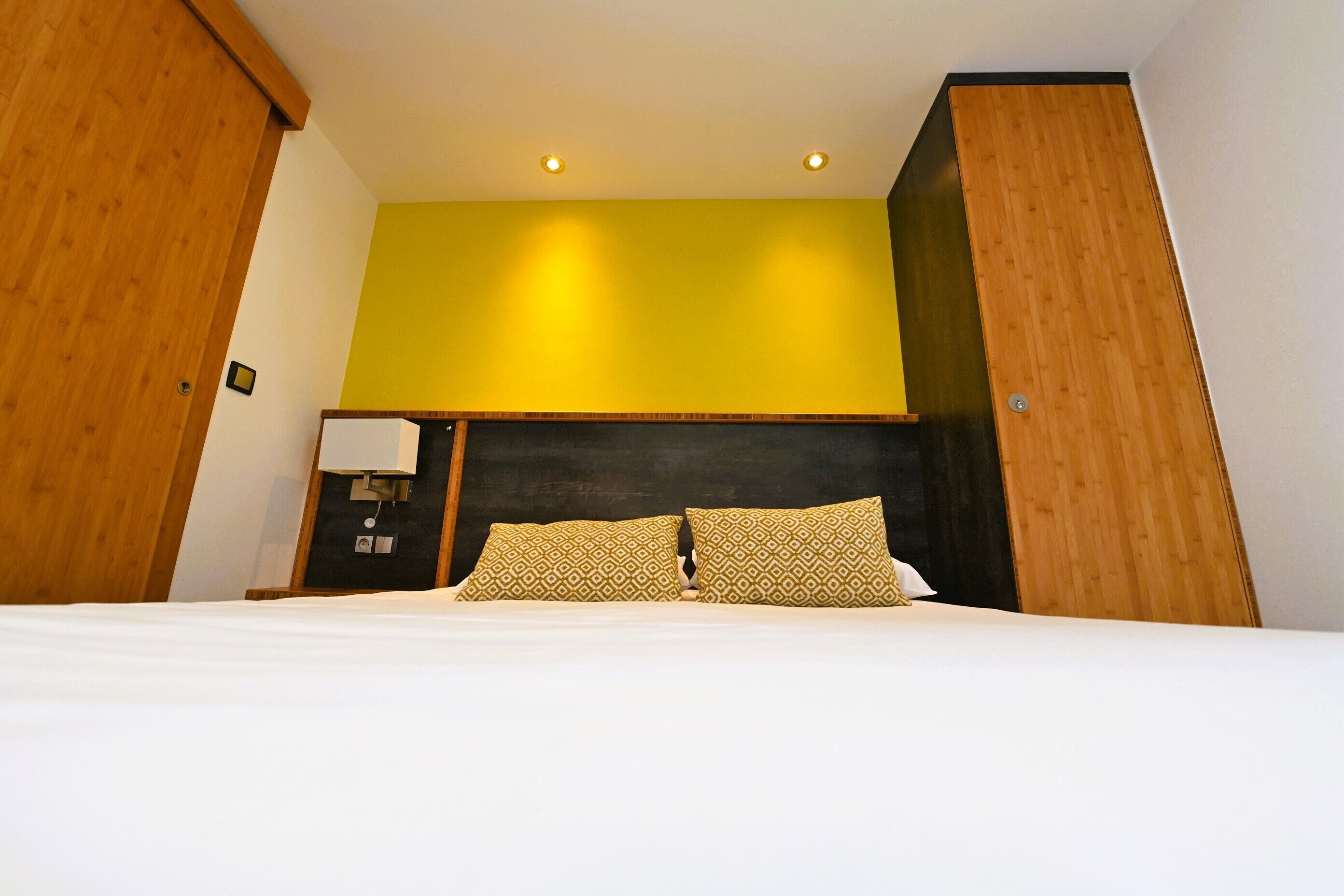 Habitación de hotel con cama, cabecera de madera, pared de acento amarilla y mesita de noche con lámpara.