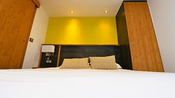 Habitación de hotel con cama, cabecera de madera, pared de acento amarilla y mesita de noche con lámpara.