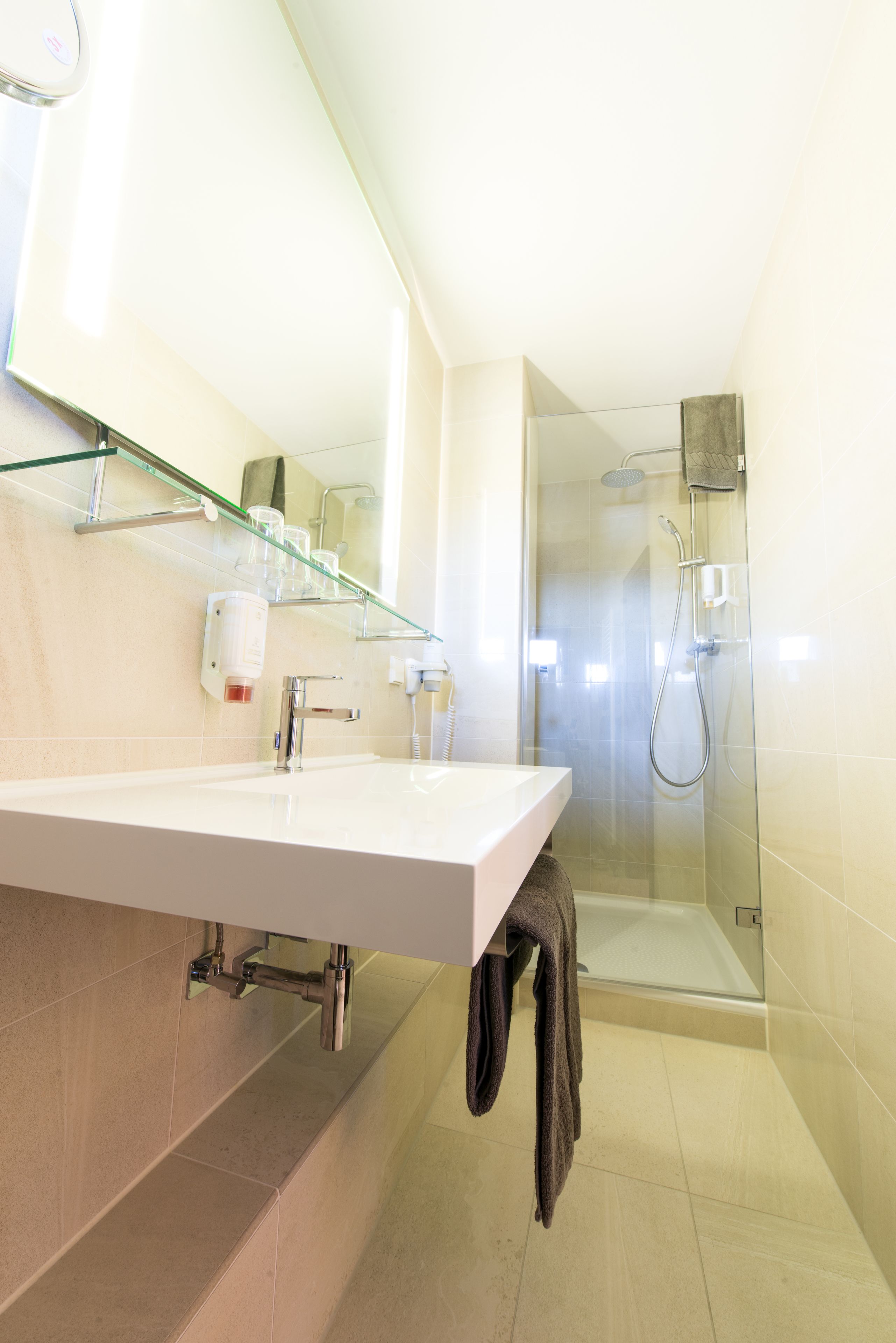Casa de banho moderna com cabine de chuveiro em vidro, lavatório branco e prateleira de vidro com produtos de higiene pessoal.