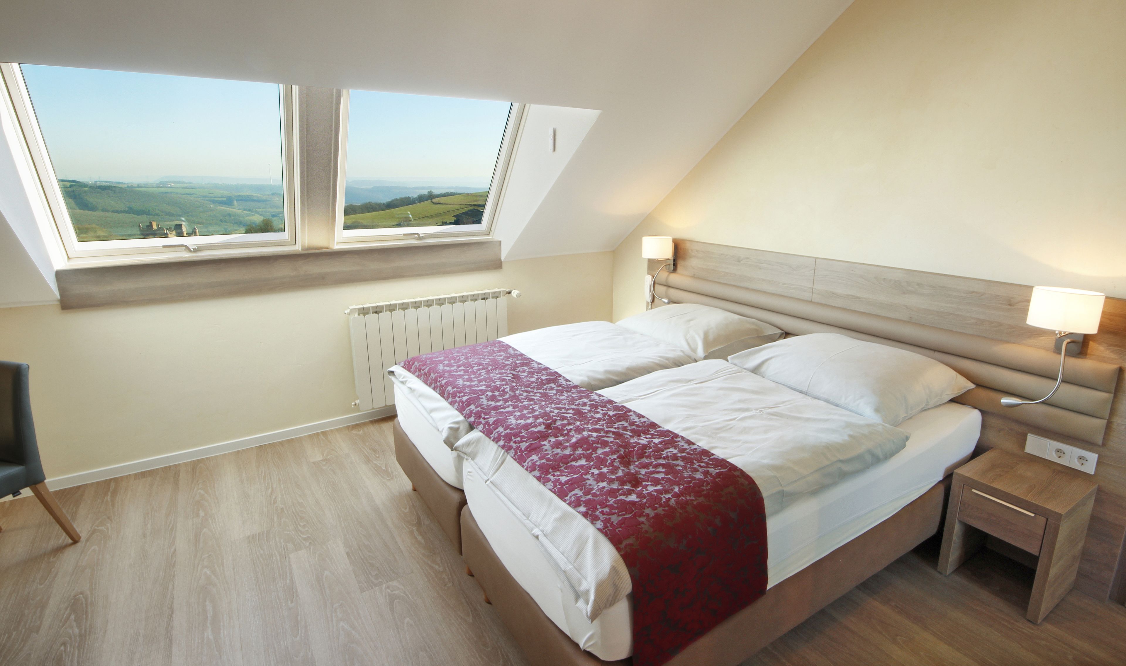 A bedroom with a bed, two windows, a bedside table, a lamp, and a view of the countryside.