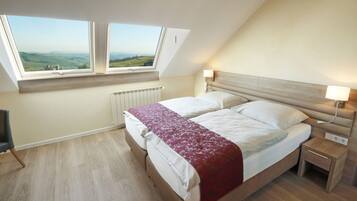 A bedroom with a bed, two windows, a bedside table, a lamp, and a view of the countryside.