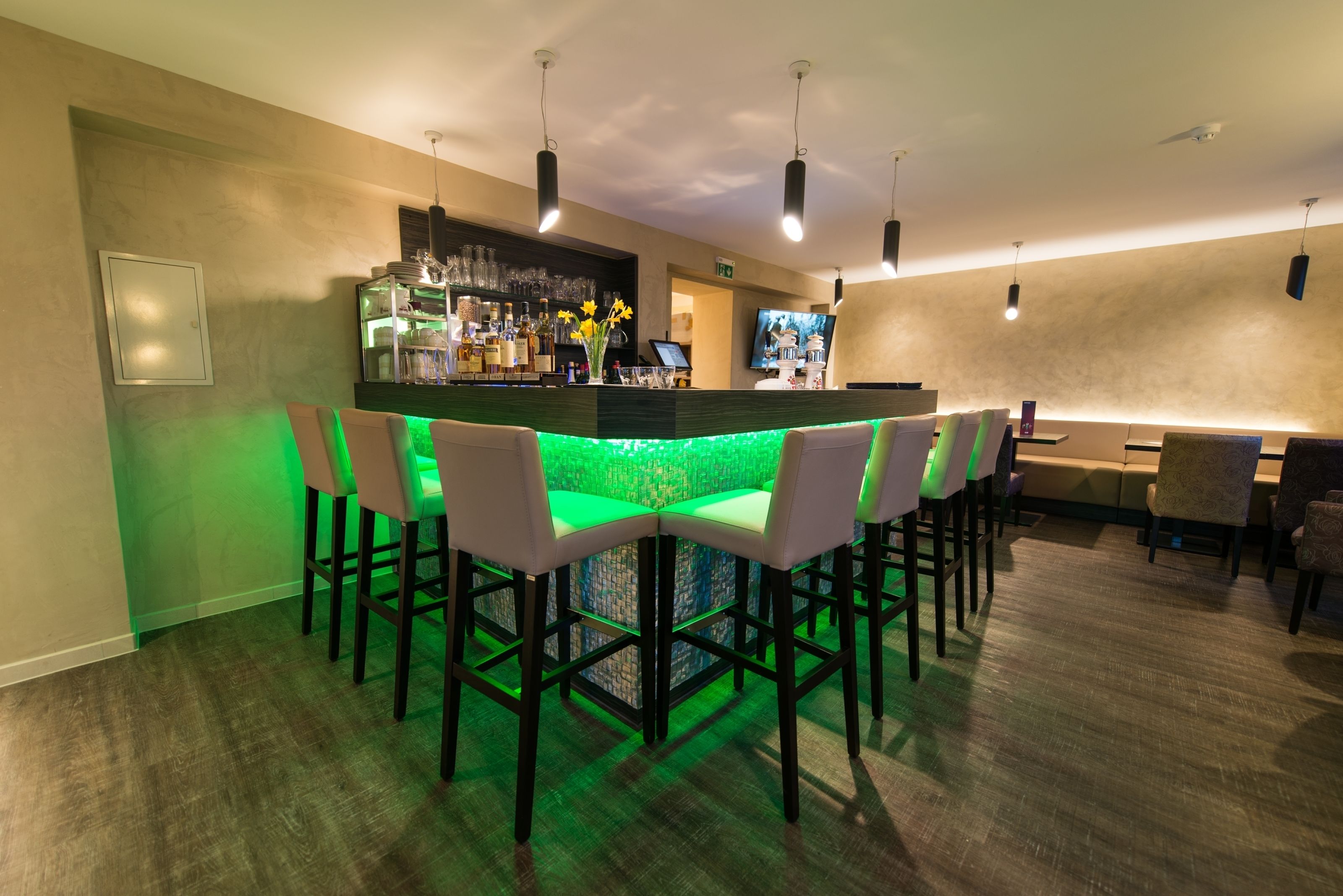 A bar area with a green-lit bar counter, white bar stools, and a television screen.