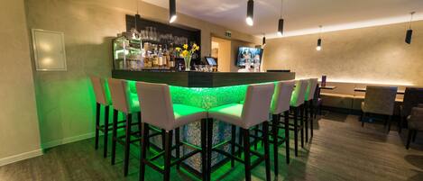 A bar area with a green-lit bar counter, white bar stools, and a television screen.