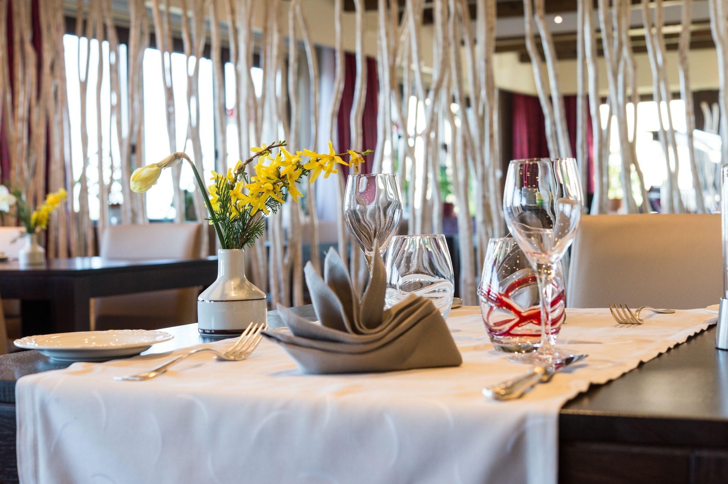 Una mesa de comedor adornada con un florero de flores amarillas, copas de vino y cubiertos.
