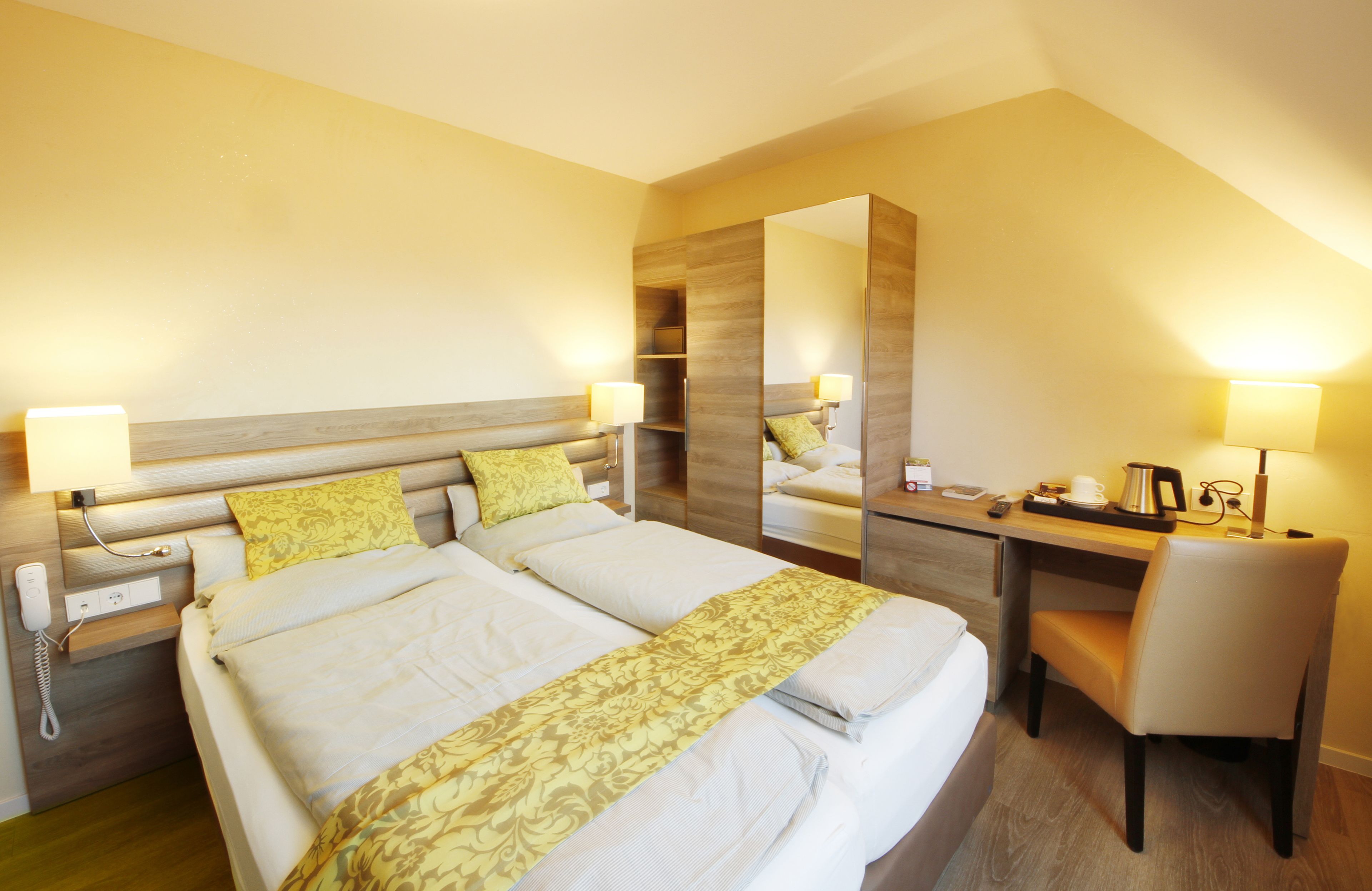 A hotel room with a large bed, a desk, and a chair. There is a mirror on the wall, a lamp on the desk, and a phone on the nightstand.