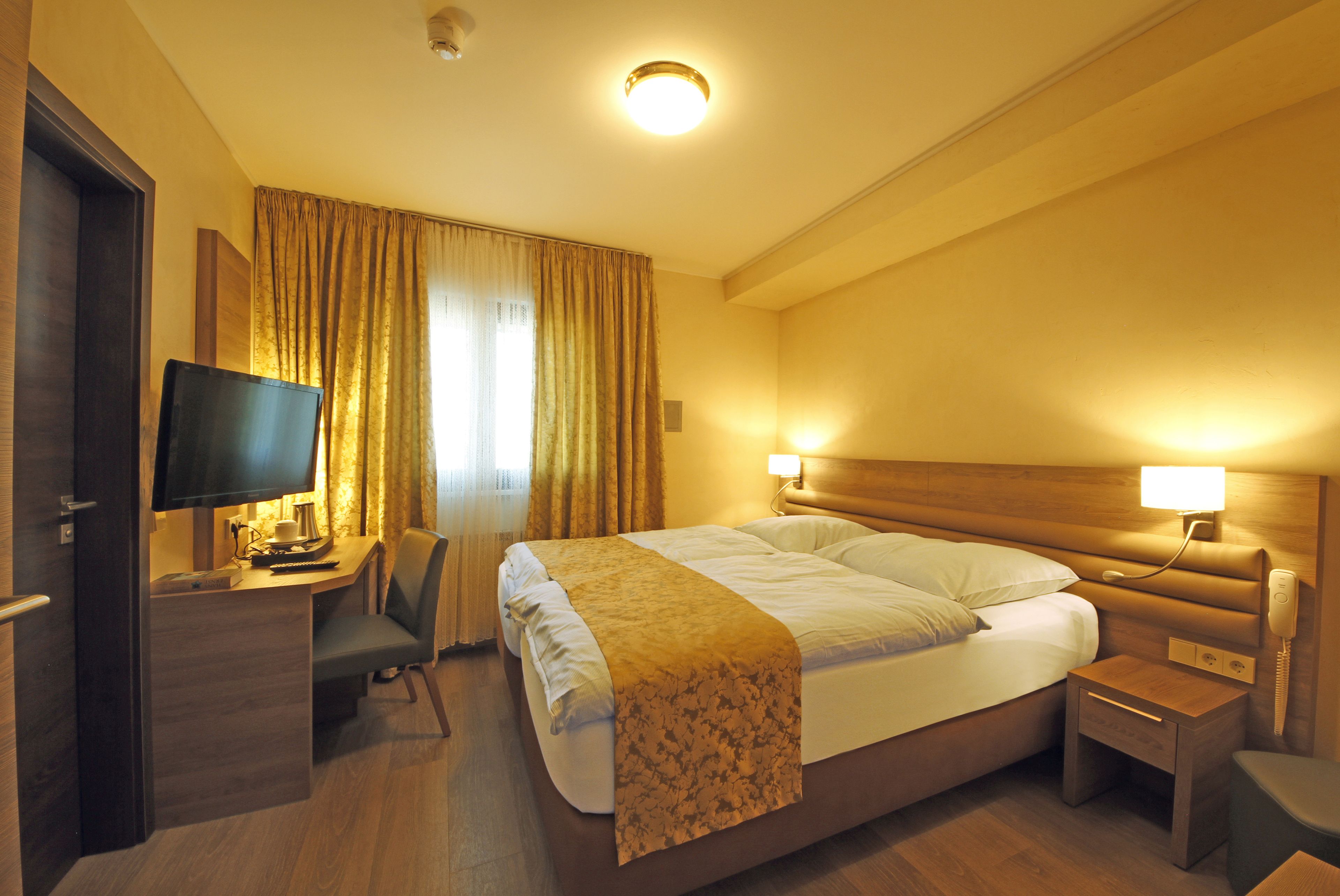 Una camera d'albergo con un letto, un comodino, una lampada, una scrivania con un computer e una TV.