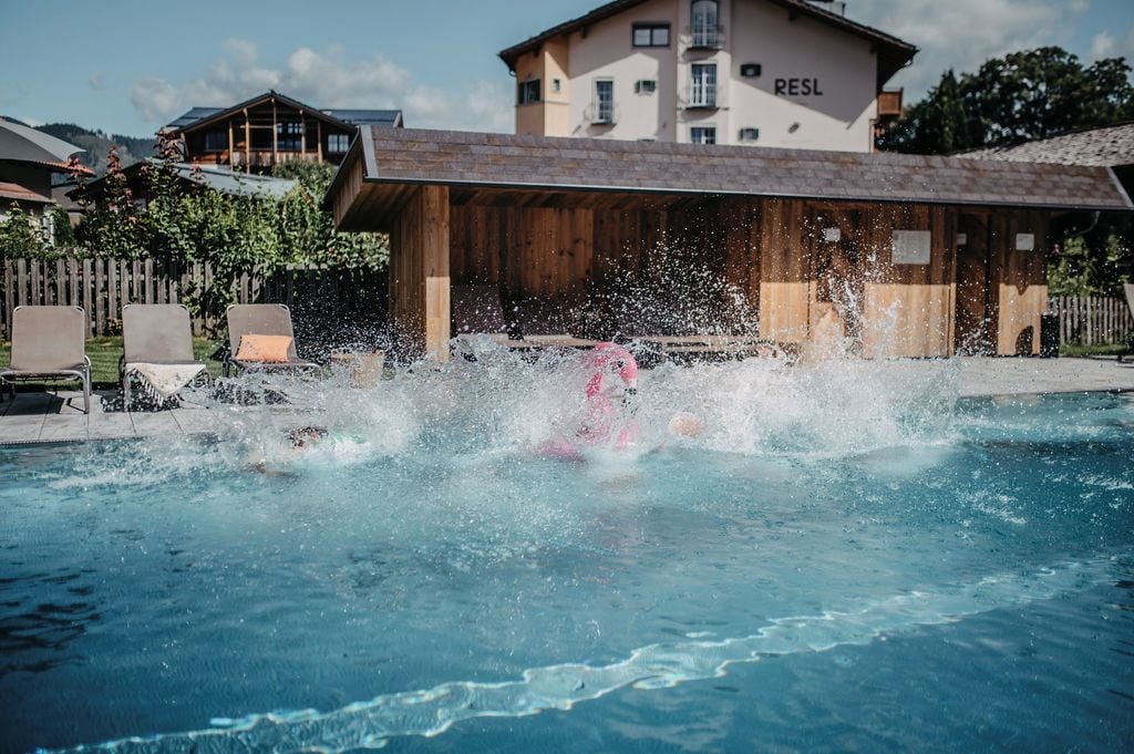 Ein Schwimmbecken mit einer Person, die ins Wasser springt, umgeben von Holzbauten und einem Gebäude im Hintergrund mit der Beschriftung 'RESL'.