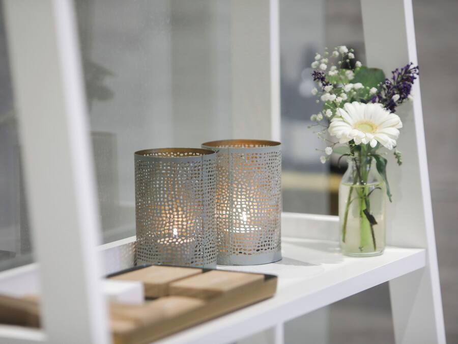 A white shelf with two perforated metal candle holders and a vase with flowers.