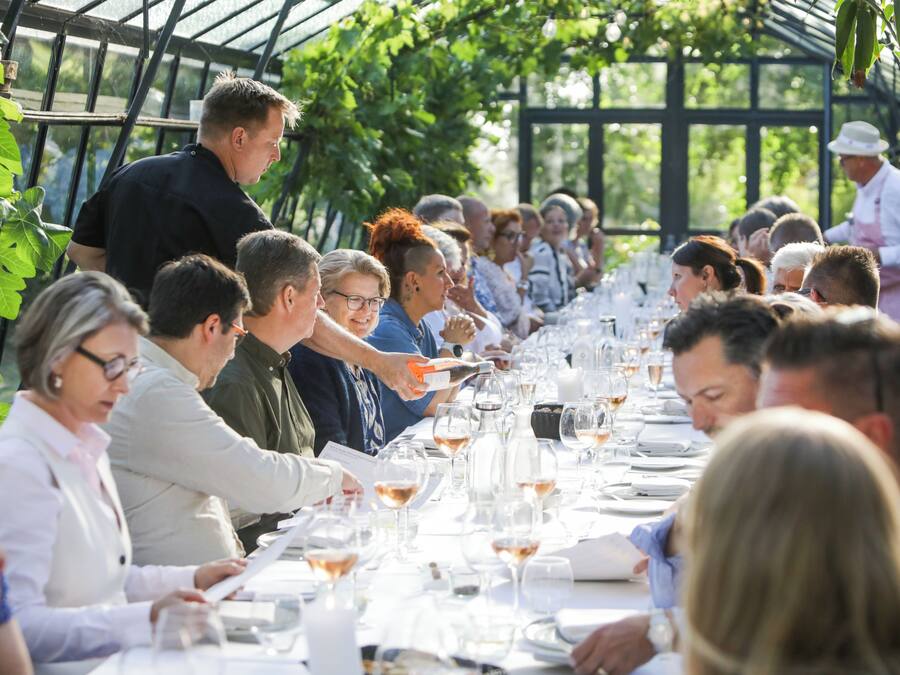 Une longue table autour de laquelle sont assis des gens, avec des verres et un serveur.