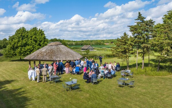 Une réunion en plein air où les participants sont assis à des tables sous une structure recouverte de chaume, entourée de pelouse et d’arbres.