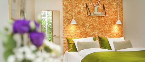 Un dormitorio con cama, almohadas, una manta verde, pared de madera y una lámpara colgante.