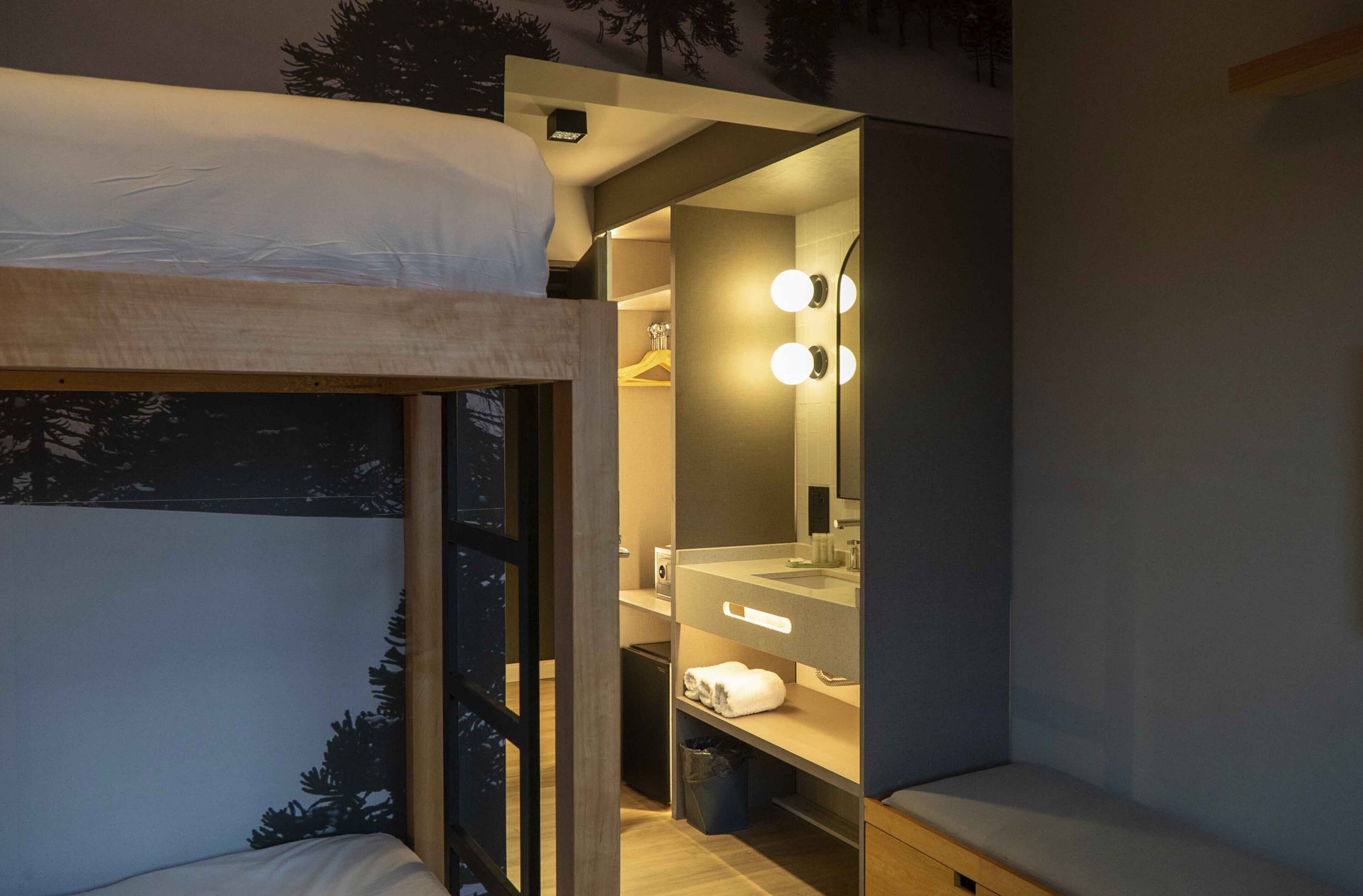 A compact bedroom with a bunk bed, a small bathroom with a mirror and lights, and a view of trees through a window.