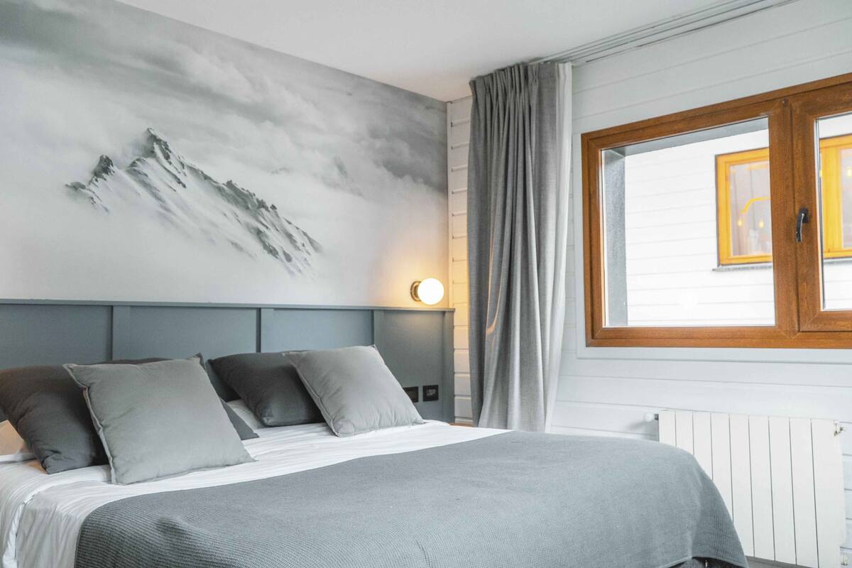 A neatly made bed with a grey bedspread and pillows, a wall-mounted light, a window with curtains, and a mountain artwork on the wall.