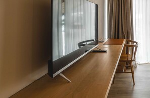 Una habitación de hotel moderna con una larga mesa de madera, un televisor de pantalla plana y un amplio ventanal con cortinas traslúcidas.