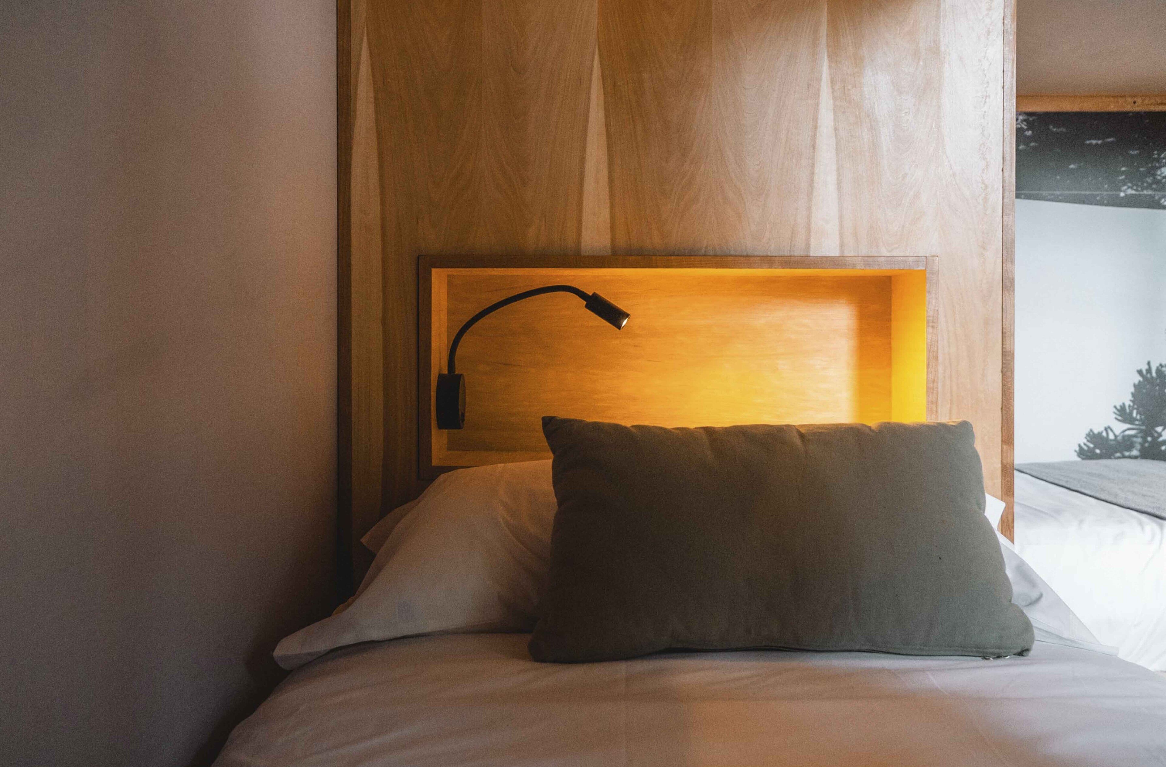 Een keurig opgemaakt bed met één kussen en een bedlampje in een houten hoofdbord.