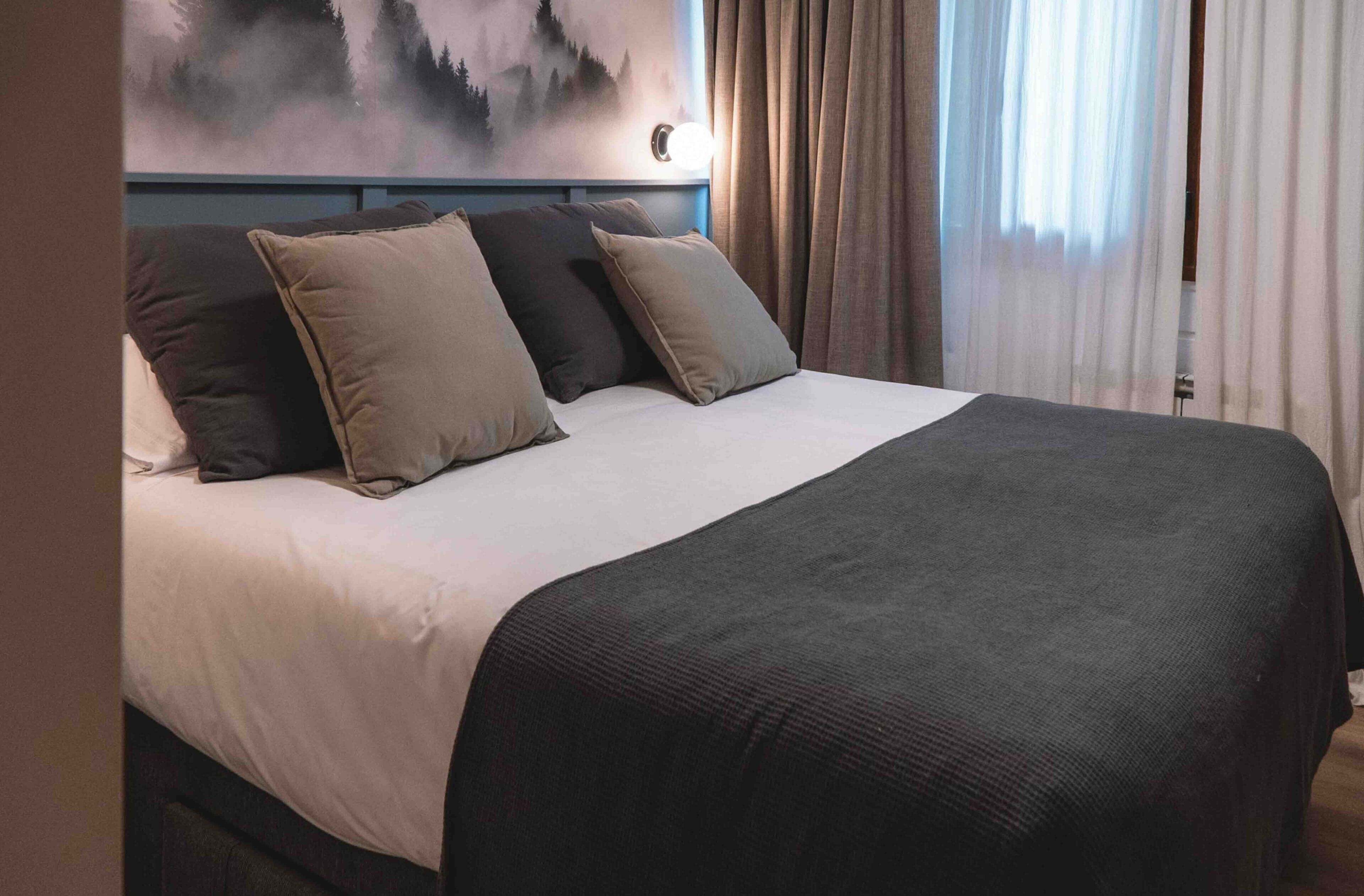 Una habitación de hotel con cama, cabecera, un cuadro y cortinas.