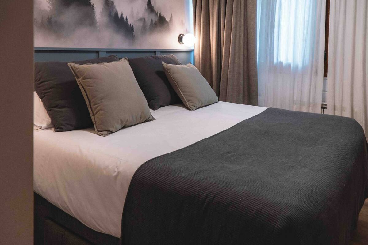 A neatly made bed with a white and dark-colored bedspread, a headboard with a painting, and a window with curtains.