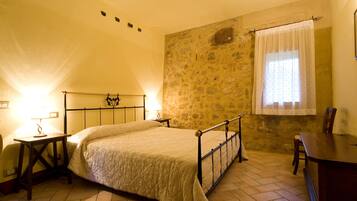 Un dormitorio con pared de piedra, una cama con estructura metálica, una mesita de noche de madera, una silla de madera y una ventana con cortina.