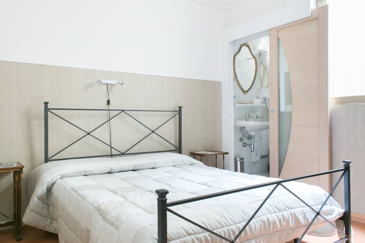 A bedroom with a bed, a bedside table, a nightstand, a mirror, and a bathroom with a sink and a towel rack.