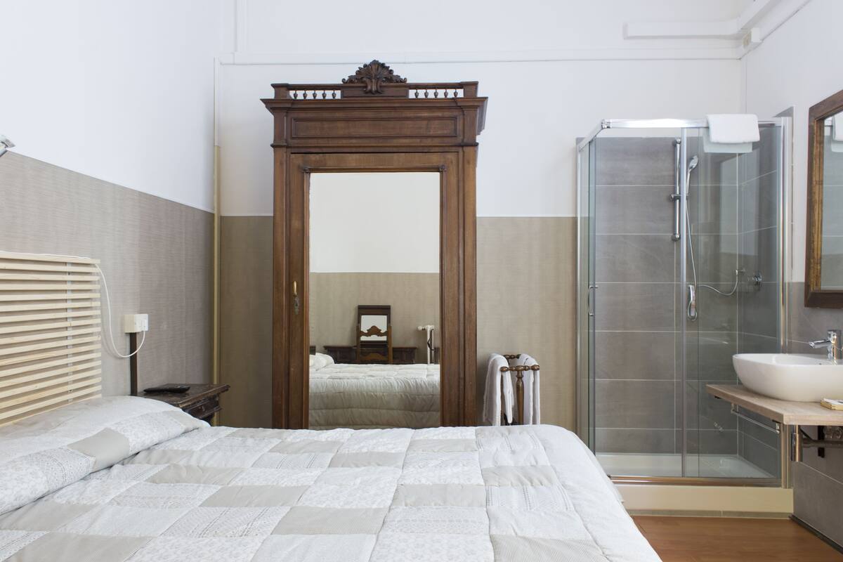 A bedroom with a large bed, a wooden wardrobe, a glass shower enclosure, and a bathroom with a sink and mirror.