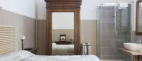 A bedroom with a large bed, a wooden wardrobe, a glass shower enclosure, and a bathroom with a sink and mirror.
