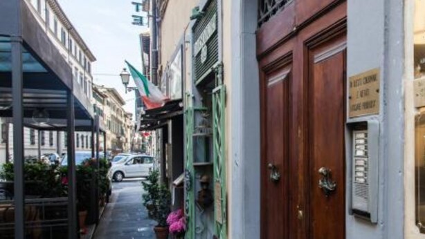 A narrow street with a building featuring a large wooden door and a sign on the wall. - Loggia Fiorentina (Florence)