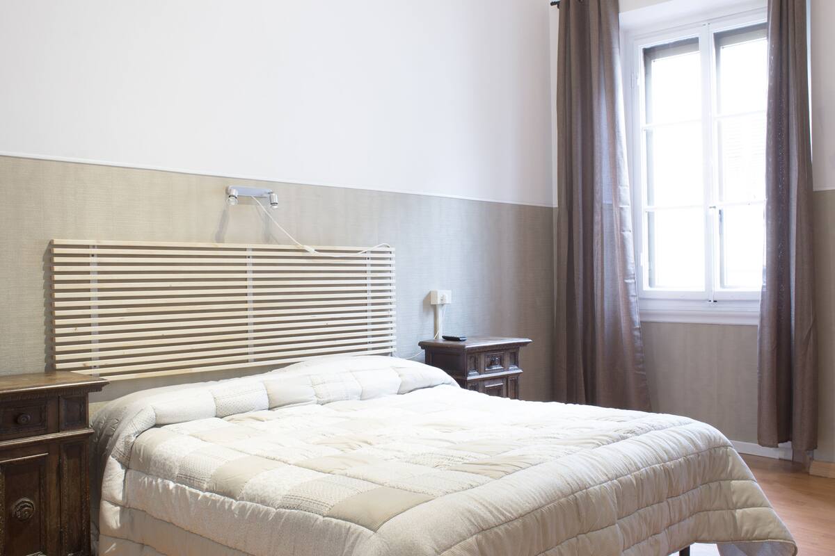 A bedroom with a bed, a nightstand, a bedside table, a window with curtains, and a headboard with a light fixture.