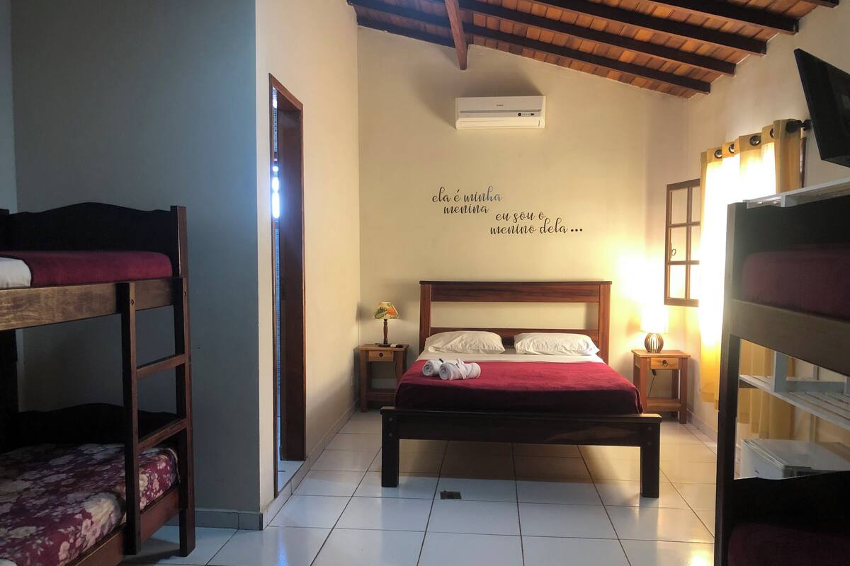 A bedroom with a wooden bunk bed, a single bed with a red blanket, a nightstand with a lamp, and a wall-mounted air conditioner.