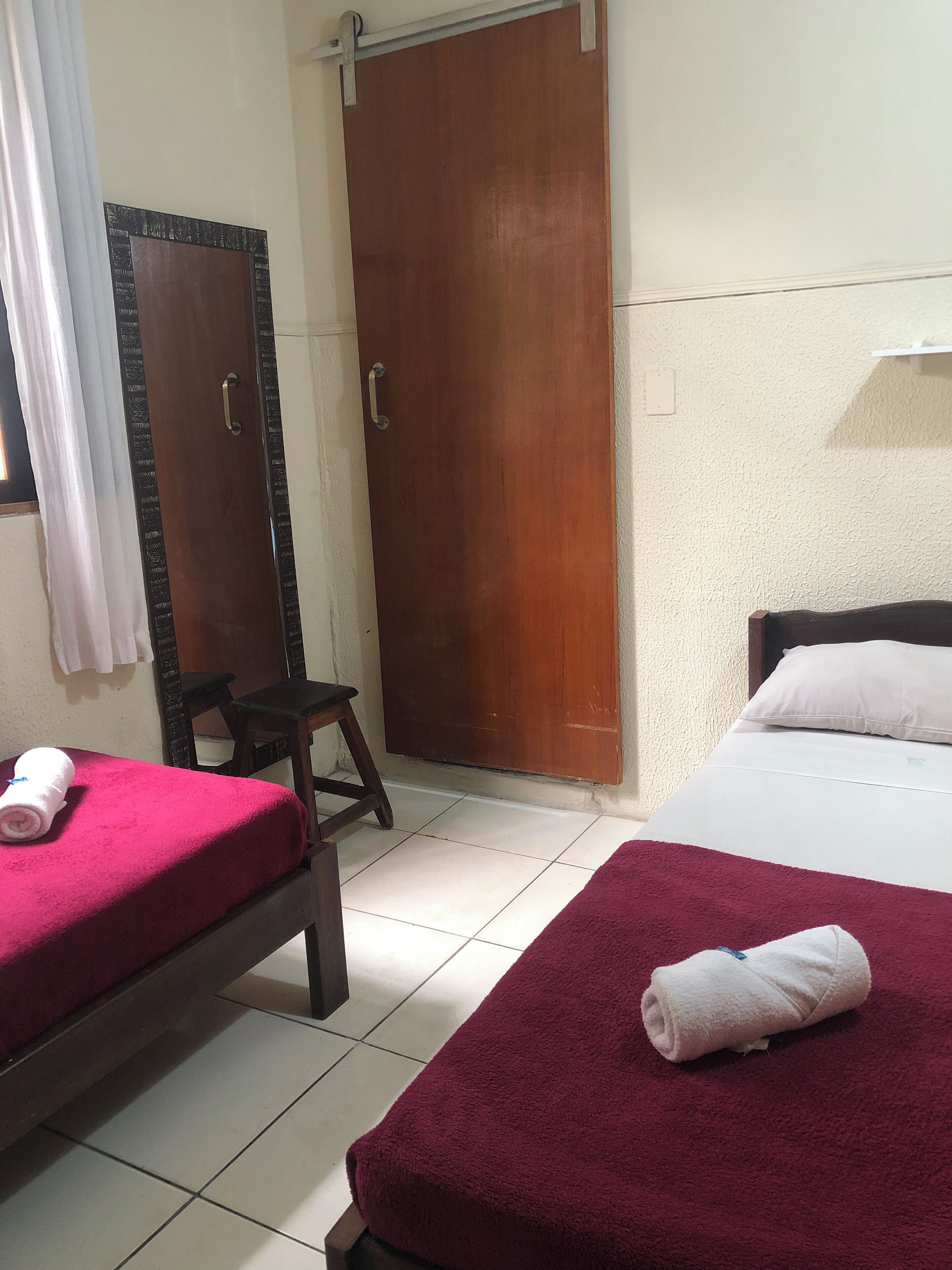 Quarto com duas camas, um guarda-roupa de madeira, um banquinho pequeno e um espelho.