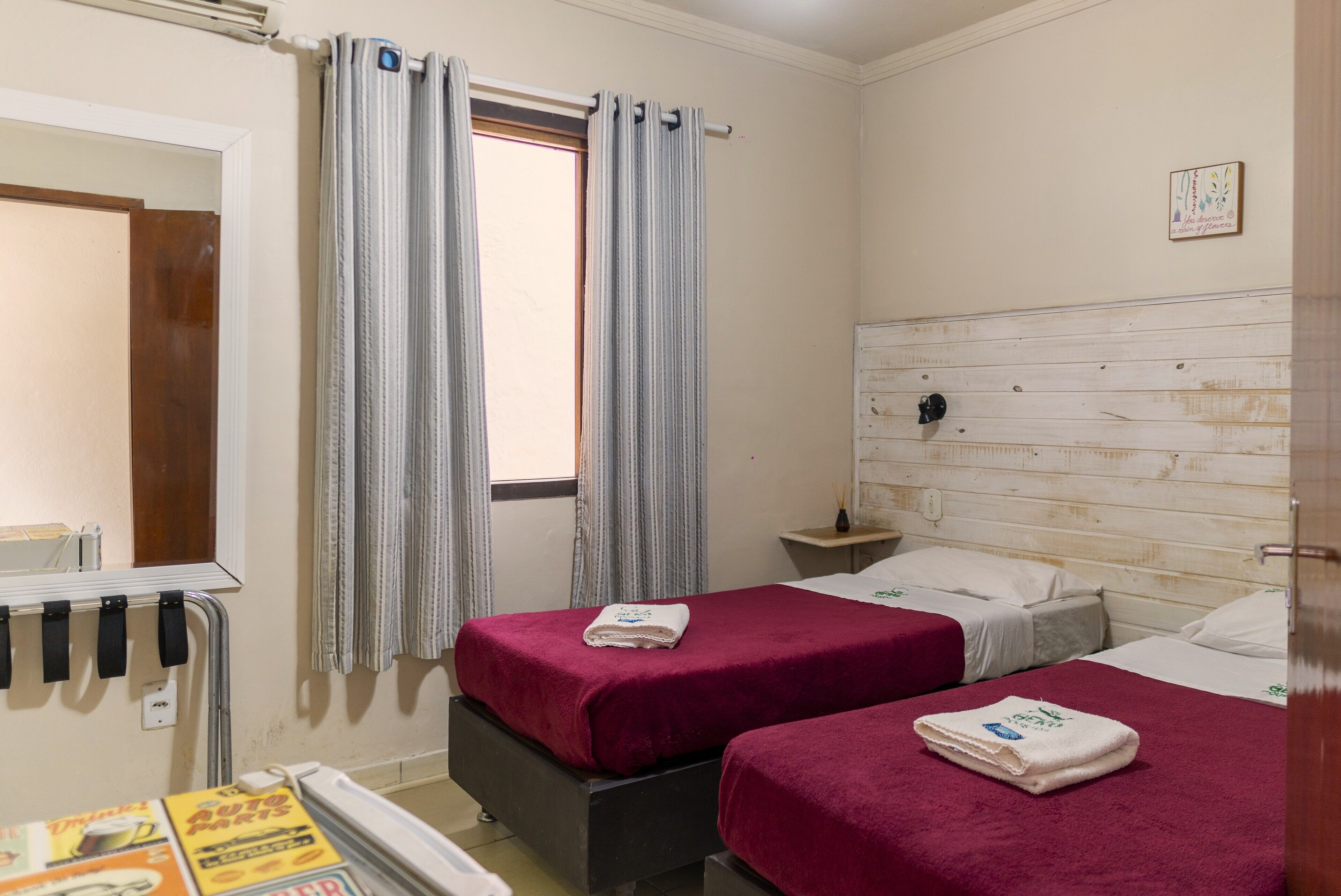 Ein Zimmer mit zwei Betten, jeweils mit roter Bettdecke und weißem Handtuch, einem hölzernen Kopfstutz, einem kleinen Nachttisch und einem Fenster mit Vorhängen.