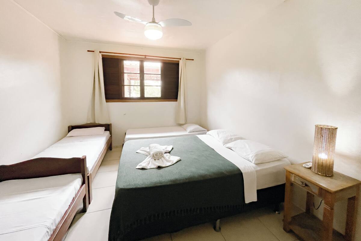 A room with two single beds, a ceiling fan, a window with curtains, and a bedside table with a lamp.