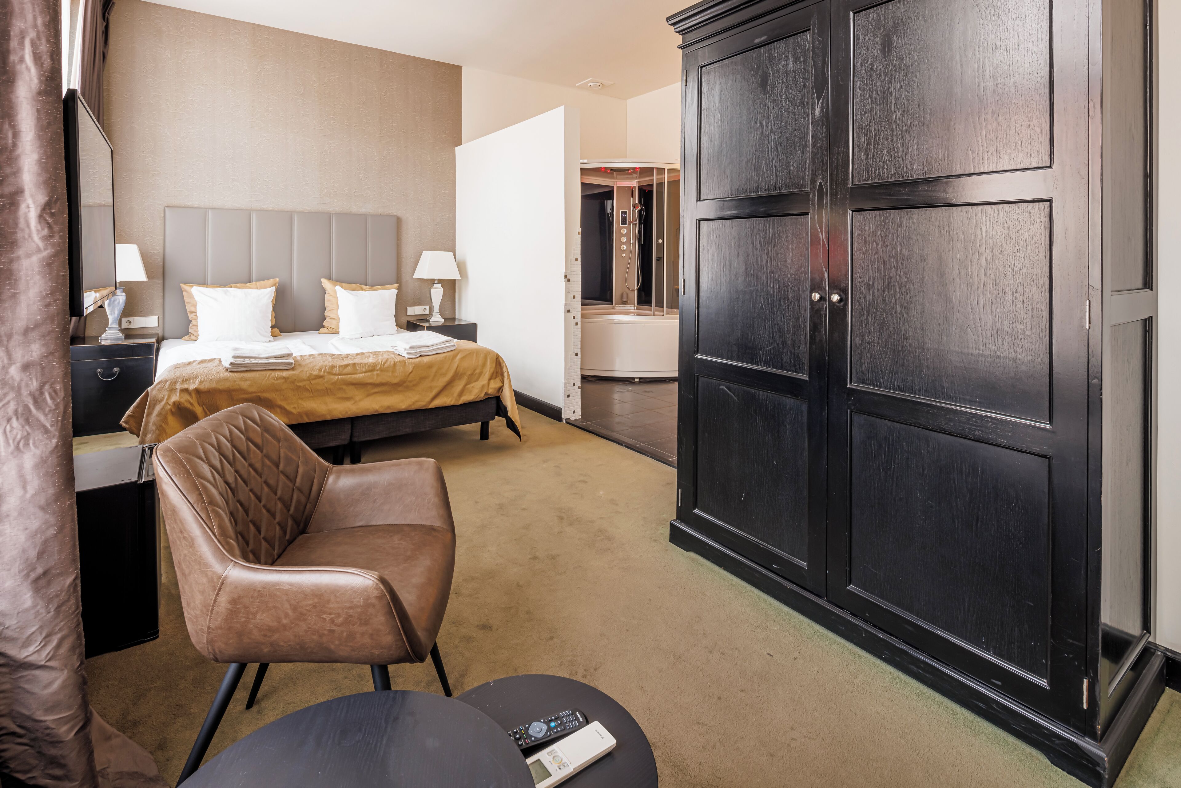 A hotel room with a bed, a chair, a desk with a lamp, a wardrobe, and a bathroom visible through an open door.