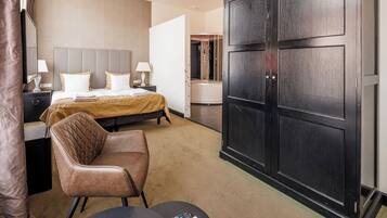 A hotel room with a bed, a chair, a desk with a lamp, a wardrobe, and a bathroom visible through an open door.
