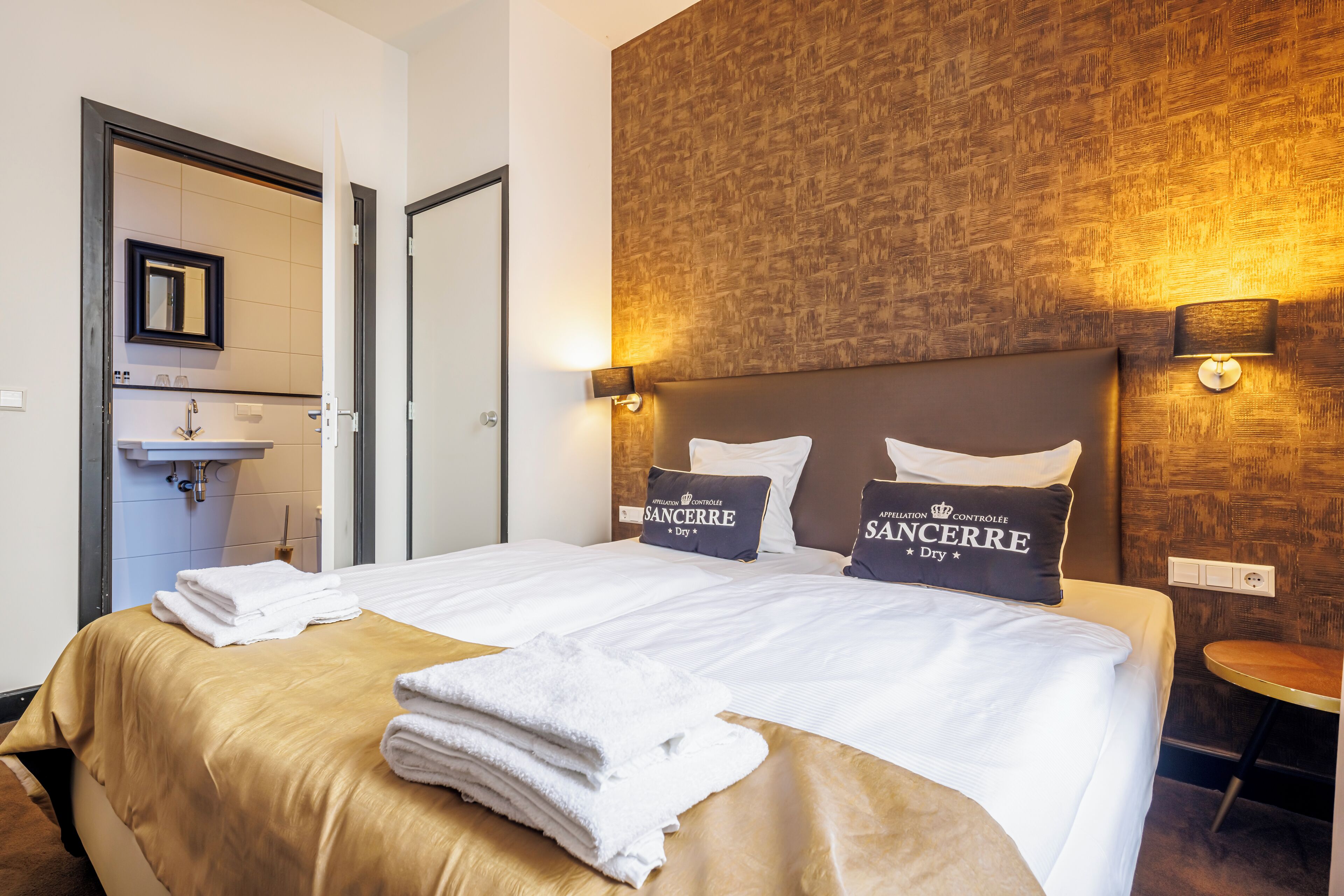 Een hotelkamer met een groot bed, twee kussens met 'SANCERRE' branding, een houten hoofdbord en een kleine ronde tafel.