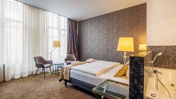 Ein Hotelzimmer mit Bett, zwei Stühlen, einem kleinen Tisch, einem Glastisch, einem großen Fenster mit durchsichtigen Vorhängen, einer Wandlampe und einem Badezimmer mit Badewanne und Dusche.