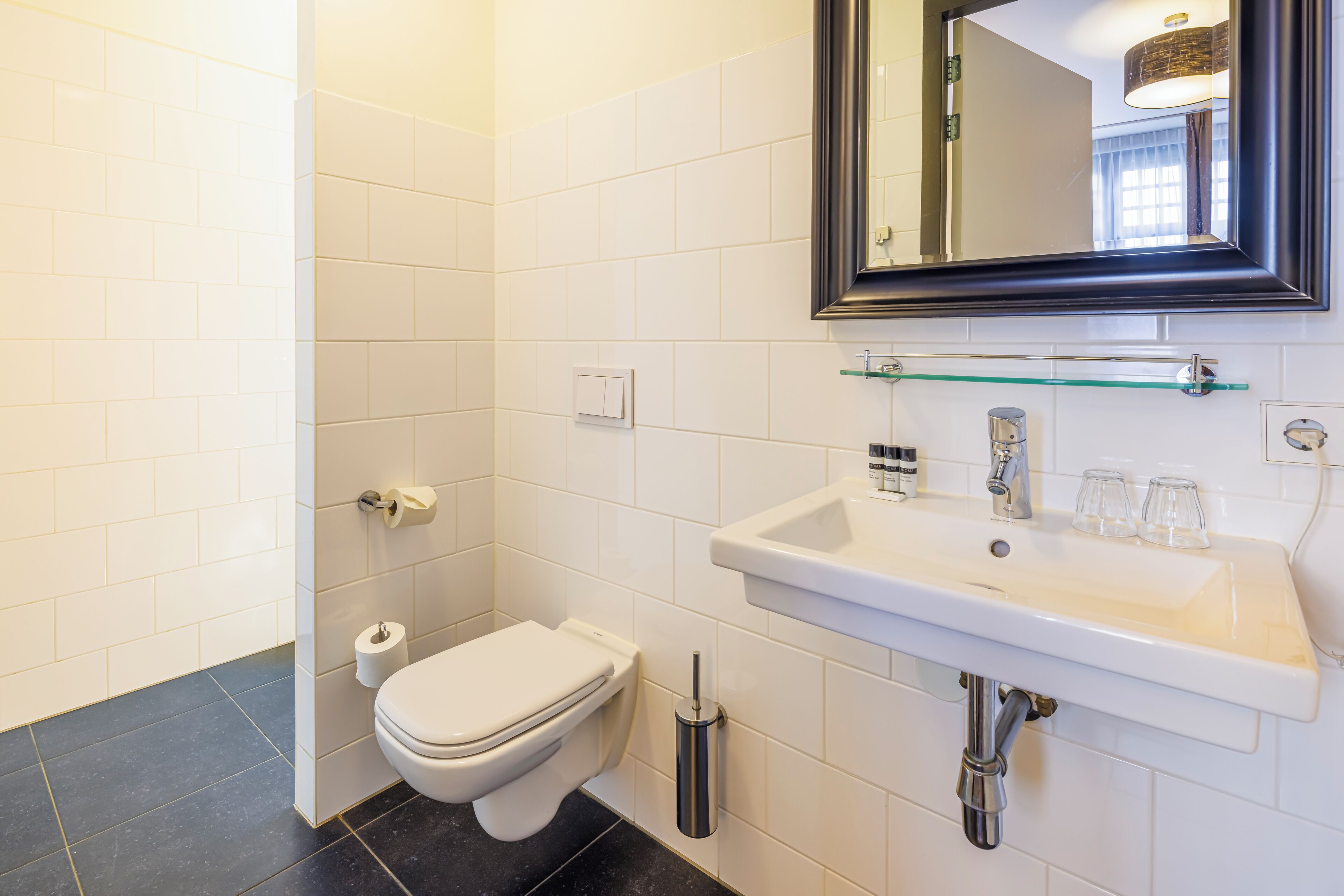 Une salle de bain moderne avec des carreaux blancs, des toilettes blanches, une étagère en verre où sont rangés les articles de toilette, et un grand miroir au-dessus du lavabo.