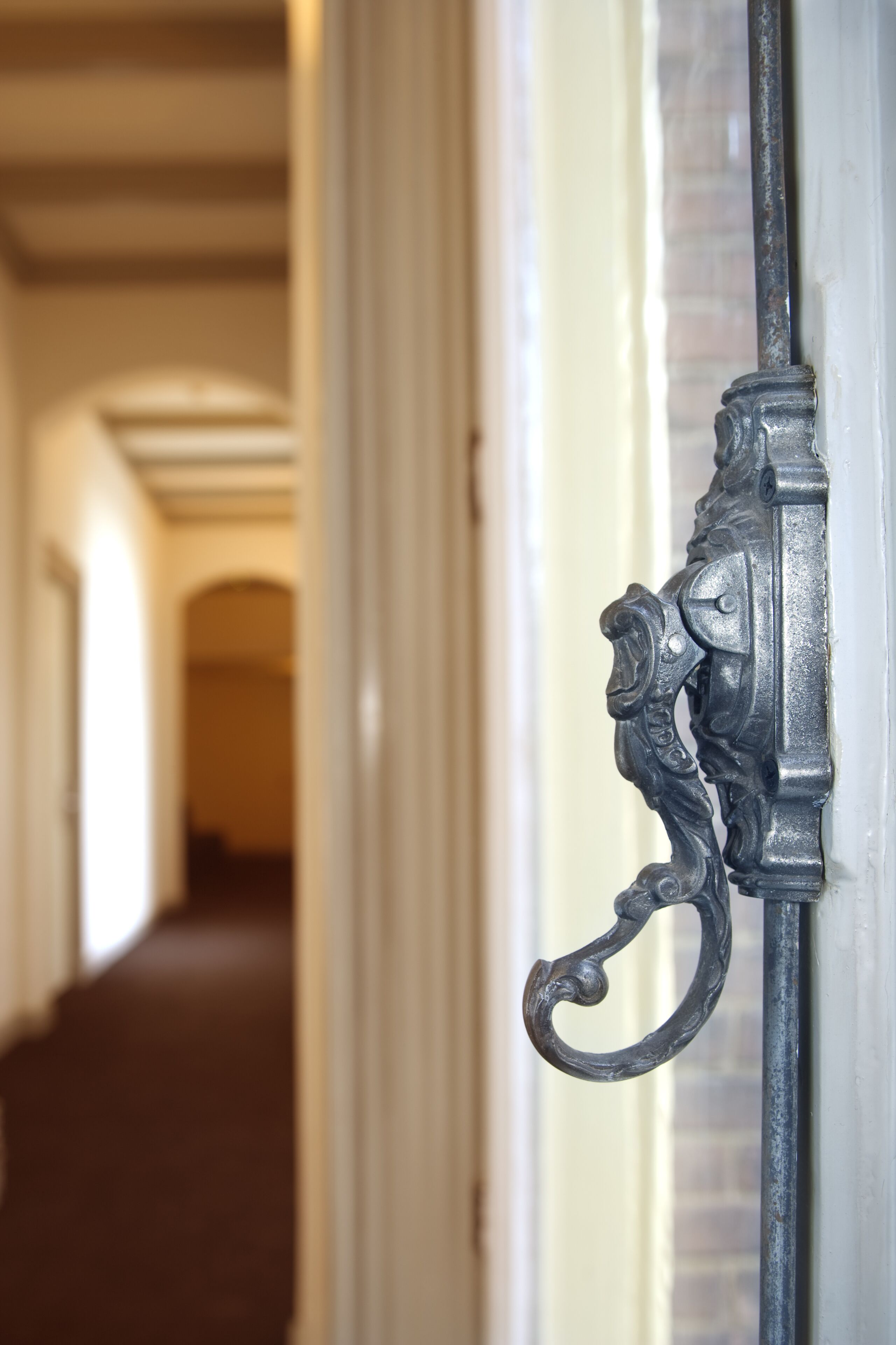 A close-up of a metal door handle with intricate design, set against a hallway with a wooden floor and white walls.