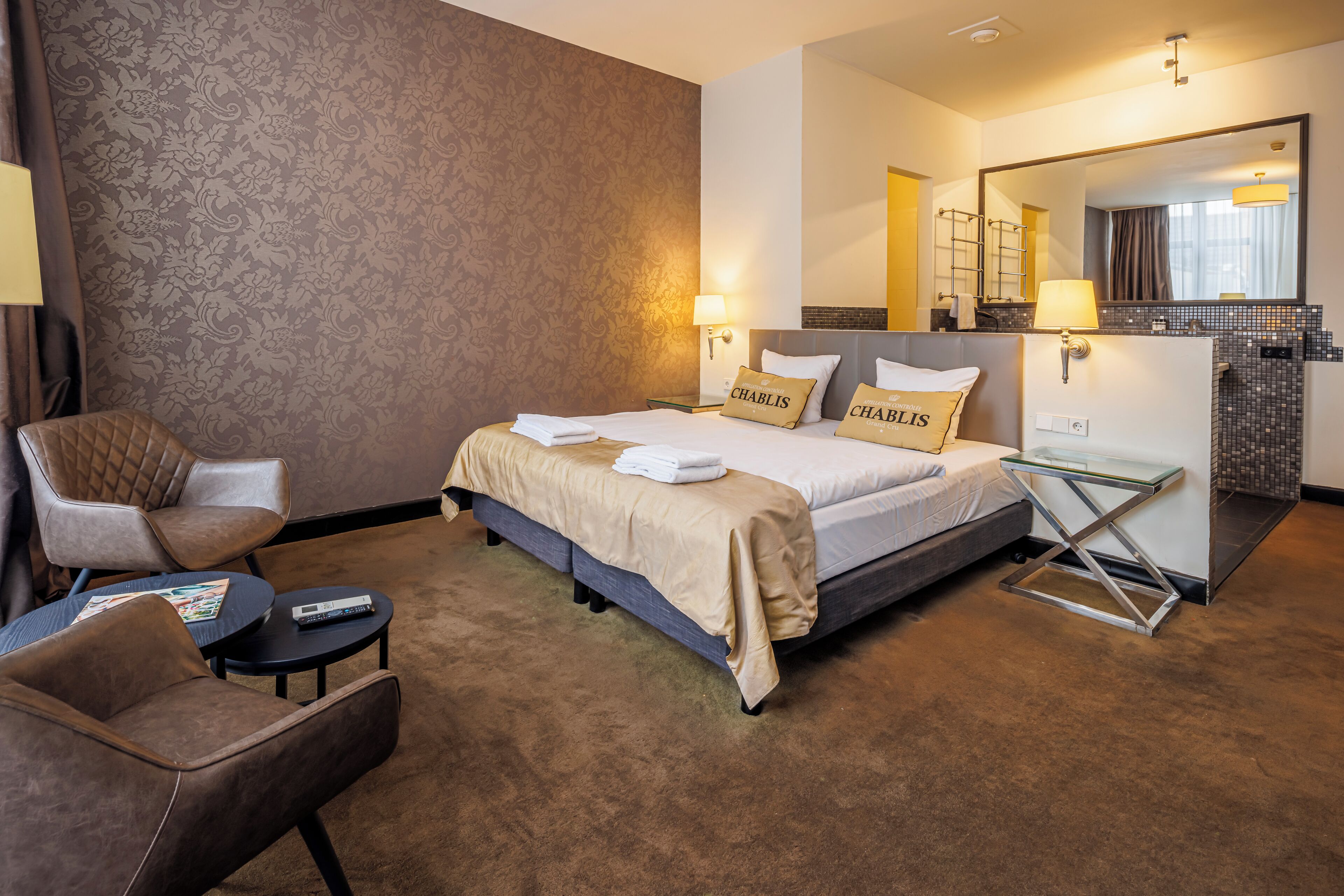 Habitación de hotel con cama, dos almohadas con el nombre 'Chablis', una silla, una mesita, una lámpara y un espejo.