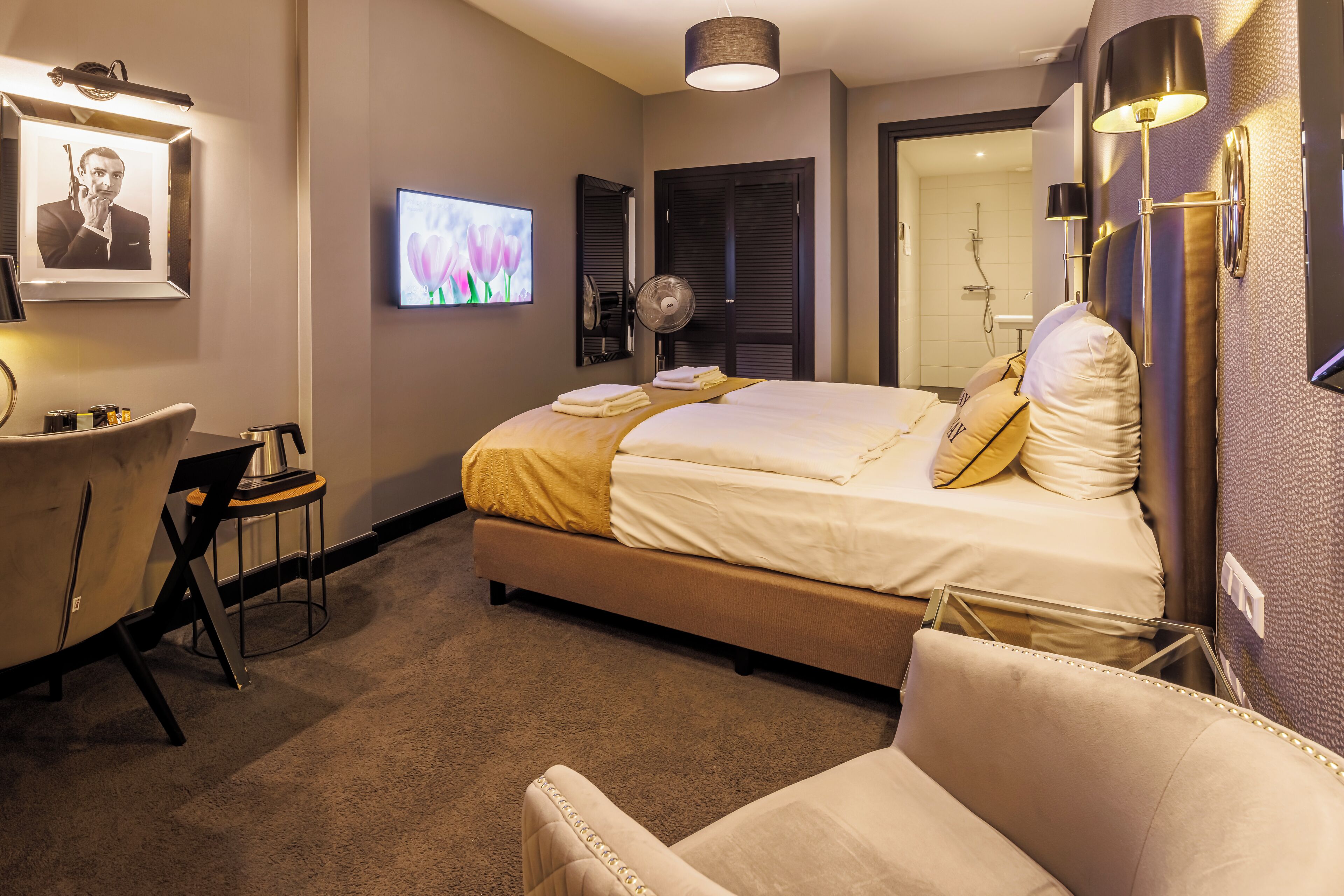 A hotel room with a bed, a desk, a chair, a TV, and a wall-mounted lamp.
