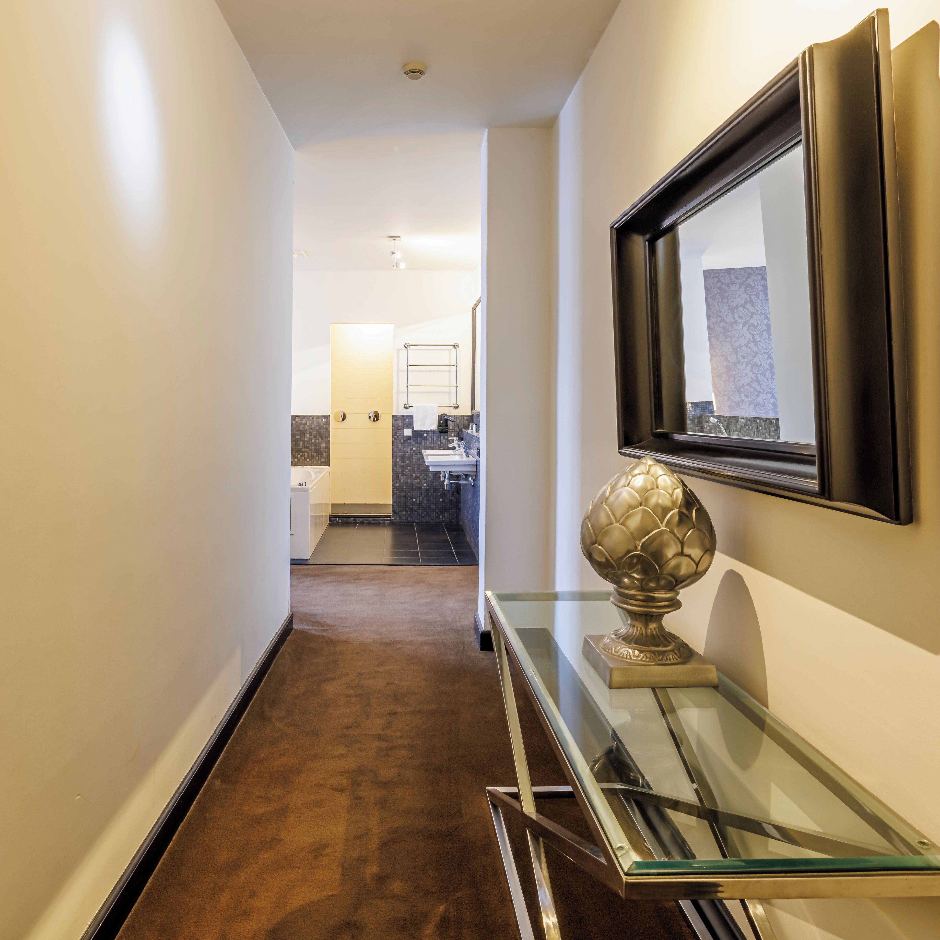 A hallway with a glass console table, a gold decorative object, and a framed mirror.