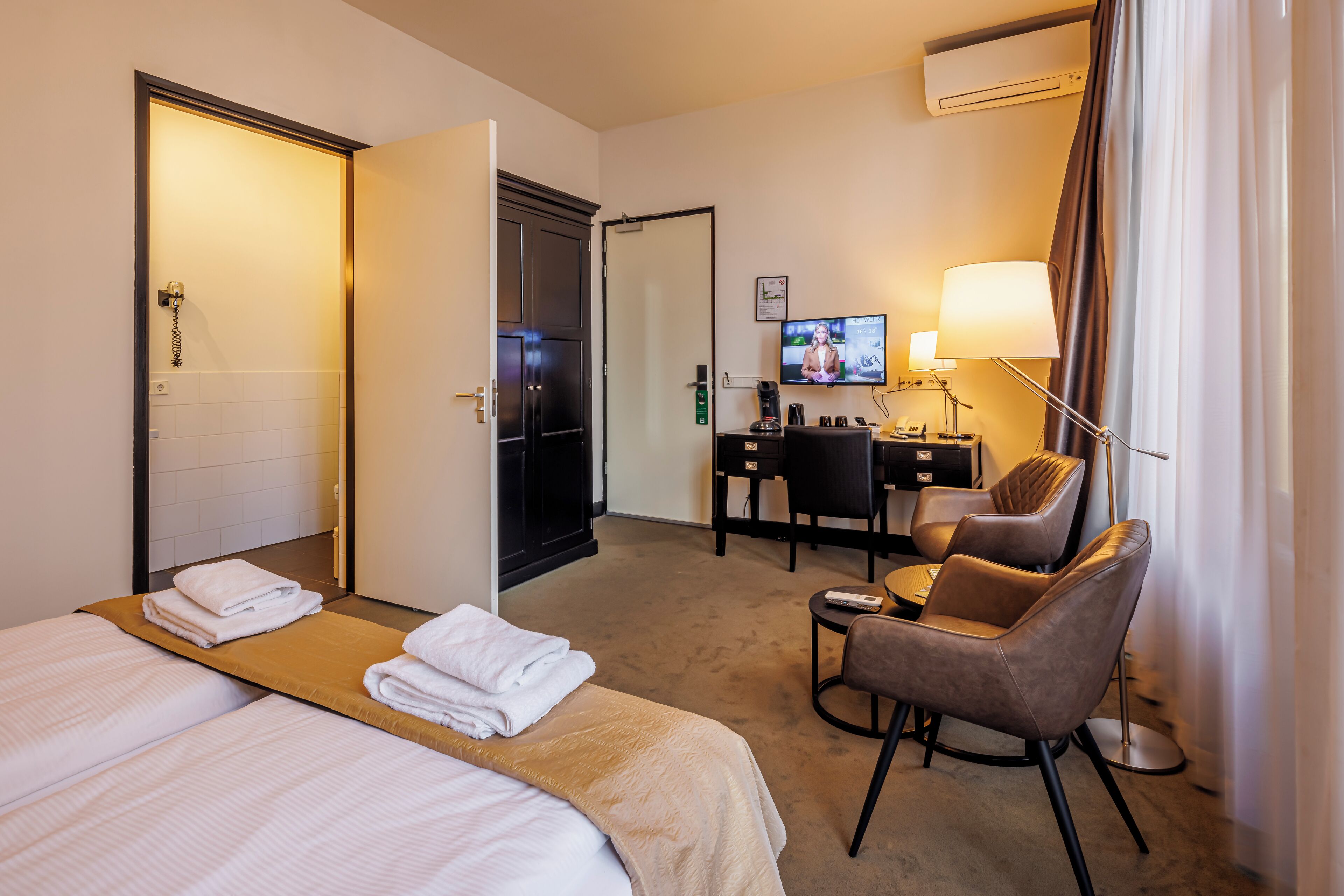 A hotel room with two beds, a desk, a chair, a TV, and a lamp.