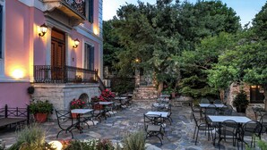 Un patio al aire libre con suelo de piedra, mesas y sillas, rodeado de exuberante vegetación y un edificio rosa con balcón.