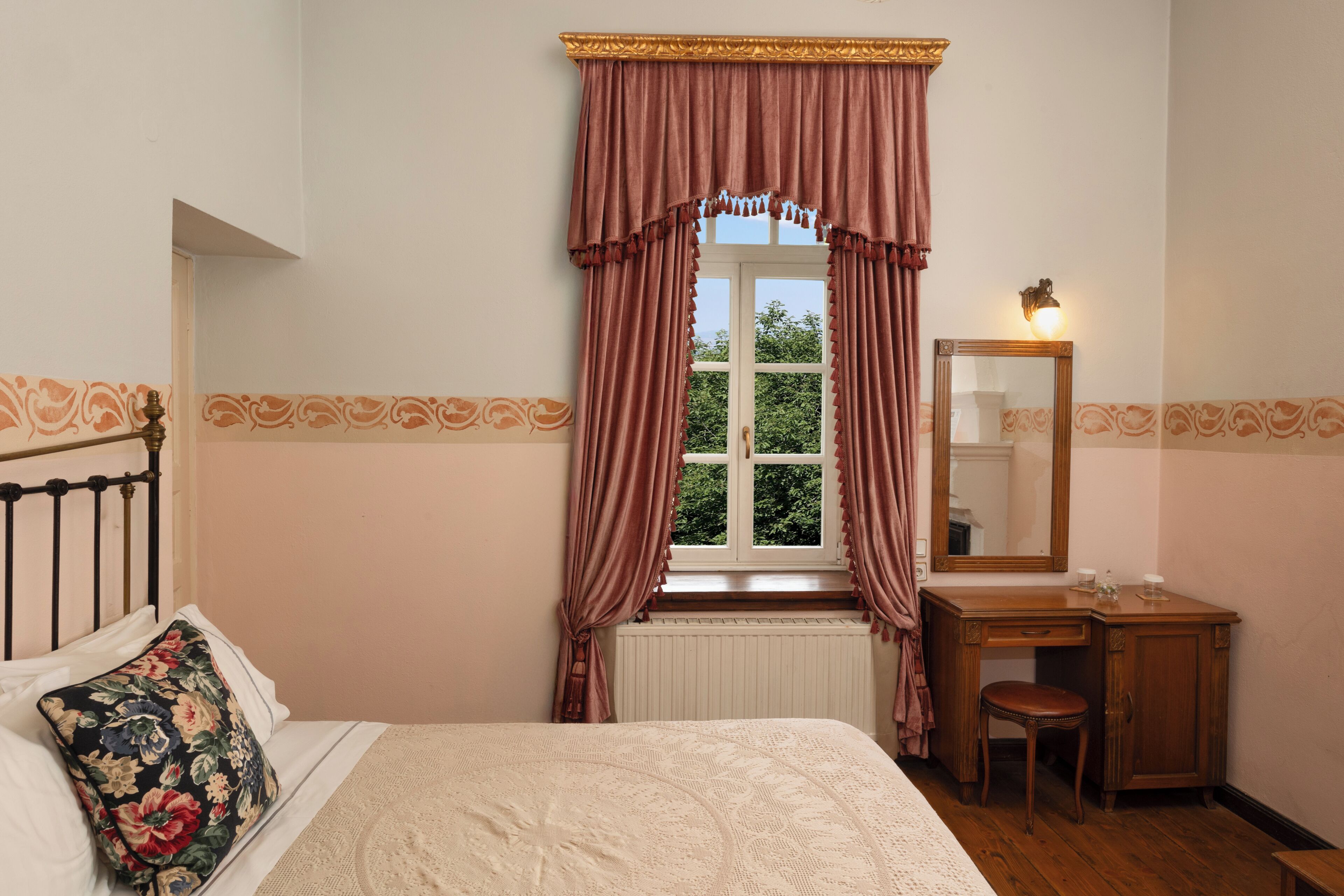A bedroom with a bed, a window with curtains, a wooden desk with a chair, and a mirror.