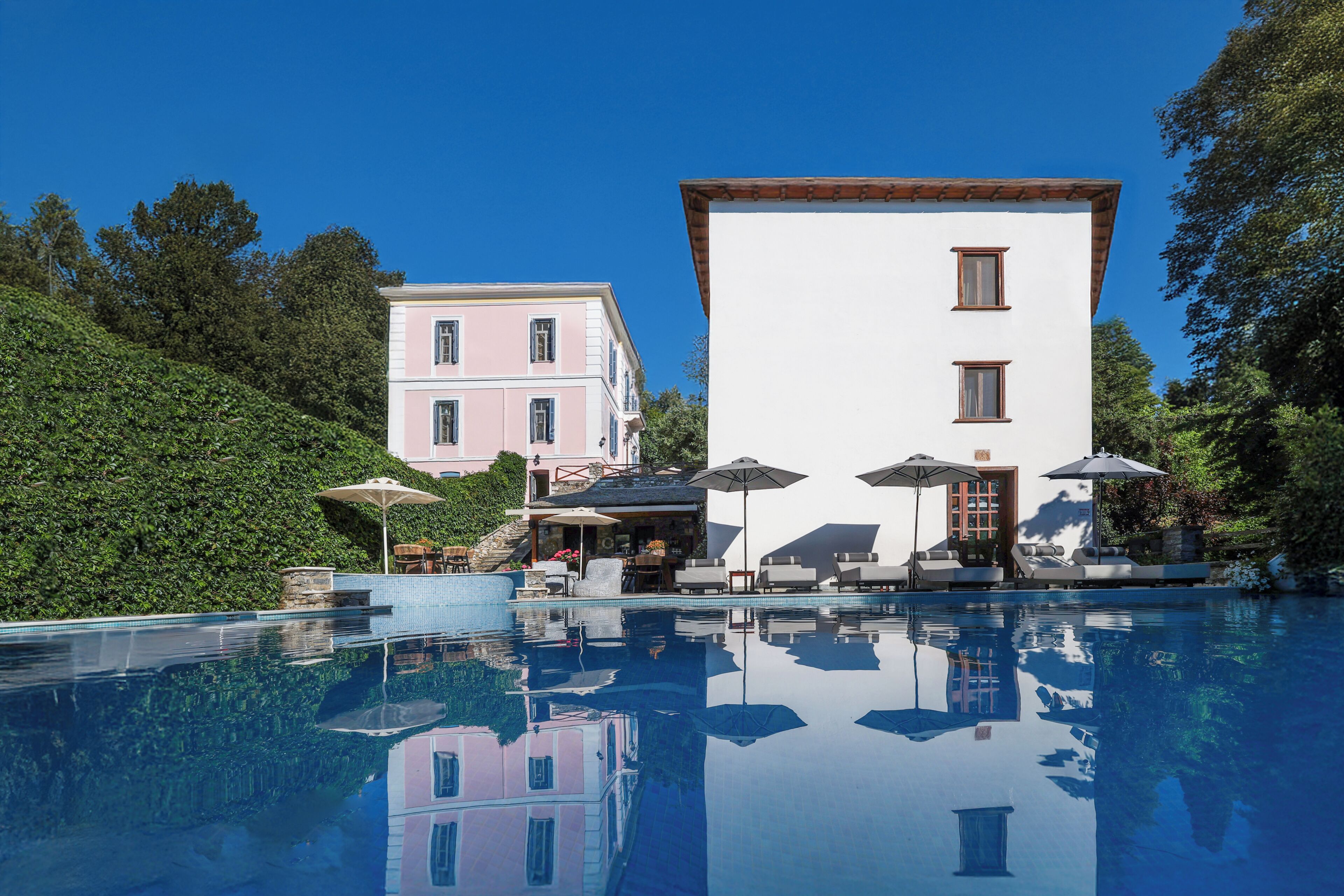 Una piscina con sillones de descanso y sombrillas, un edificio con balcón y un paisaje de colinas al fondo.