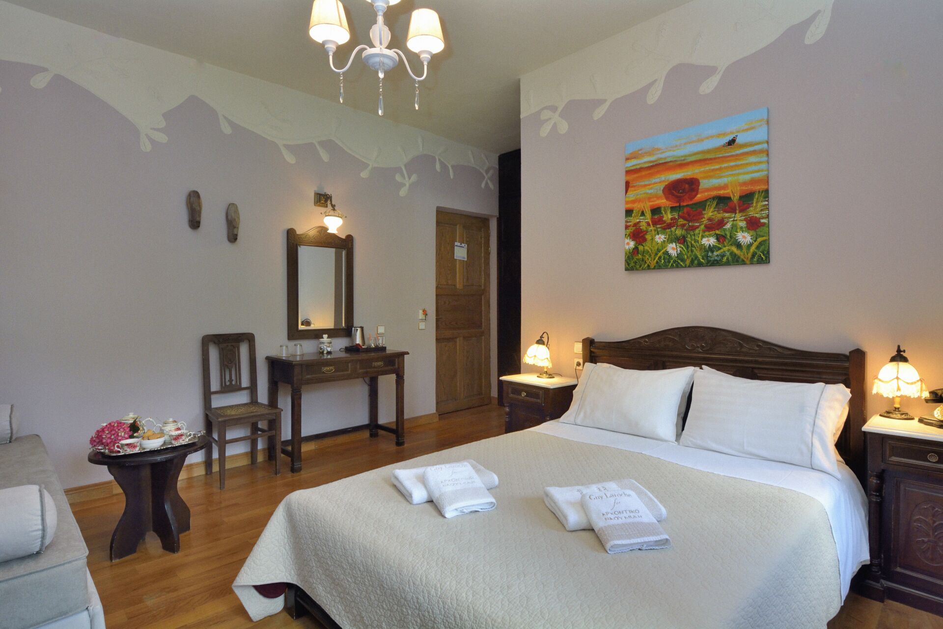 A hotel room with a bed, bedside tables, a desk, a chair, and a painting on the wall.
